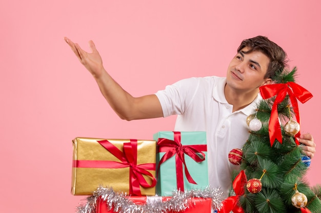 Vooraanzicht van jonge man rond kerstcadeautjes en vakantieboom op roze muur