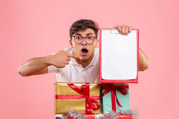 Vooraanzicht van jonge man rond kerstcadeautjes met dossiernota op de roze muur