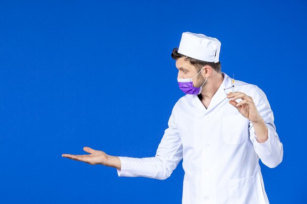 Vooraanzicht van jonge mannelijke arts in medisch kostuum en de injectie van de maskerholding op blauw