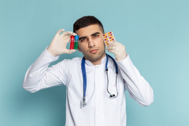 Vooraanzicht van jonge mannelijke arts in wit kostuum met blauwe de kolven en pillen van de stethoscoopholding
