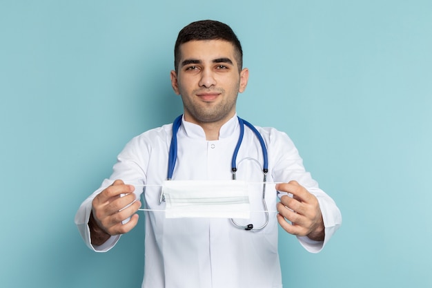 Vooraanzicht van jonge mannelijke arts in wit pak met blauwe stethoscoop glimlachend en masker te houden