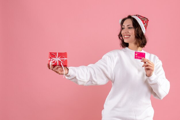 Vooraanzicht van jonge vrouw die kleine aanwezige Kerstmis en bankkaart op roze muur houdt