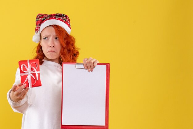 Vooraanzicht van jonge vrouw die kleine aanwezige Kerstmis houdt en nota met droevig gezicht op gele muur indient