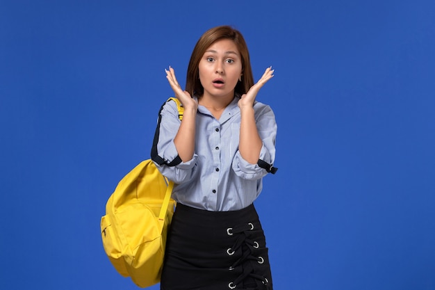 Vooraanzicht van jonge vrouw in blauw shirt met gele rugzak poseren met verbaasde uitdrukking op blauwe muur