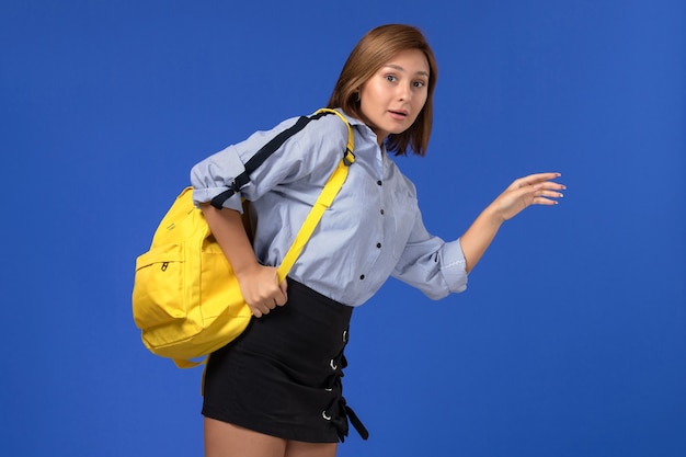 Gratis foto vooraanzicht van jonge vrouw in blauw shirt zwarte rok dragen gele rugzak poseren en draait op de lichtblauwe muur