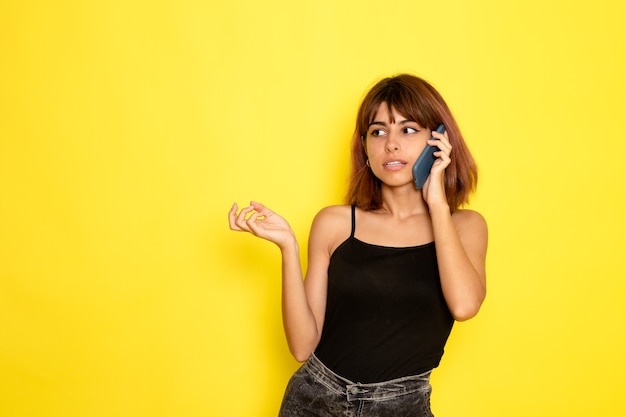 Vooraanzicht van jonge vrouw in zwart shirt en grijze spijkerbroek praten aan de telefoon op gele muur