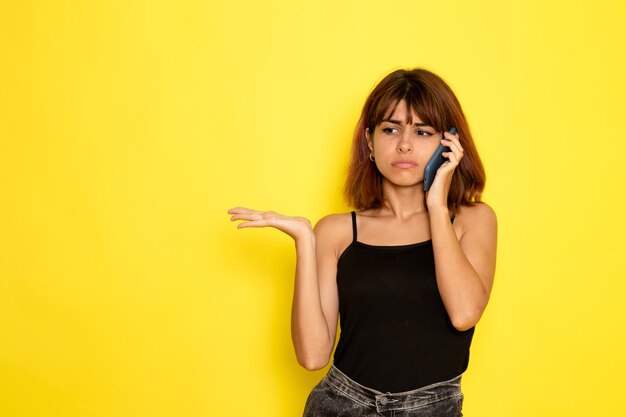 Vooraanzicht van jonge vrouw in zwart shirt praten aan de telefoon op lichtgele muur