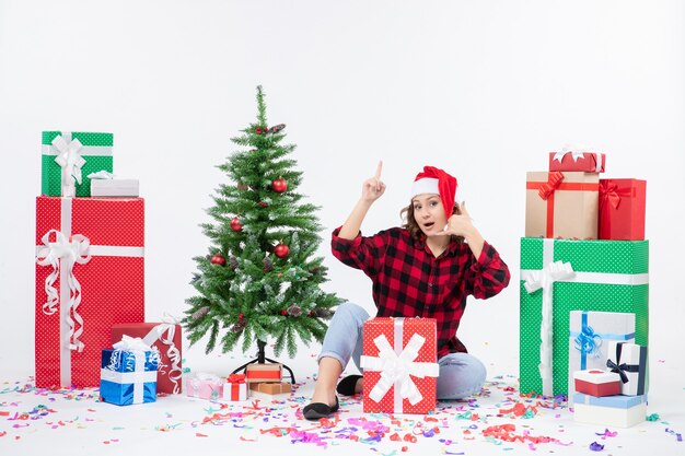 Vooraanzicht van jonge vrouw zitten rond presenteert en kleine vakantieboom op witte muur