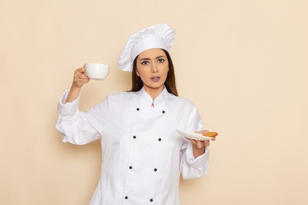 Vooraanzicht van jonge vrouwelijke kok in wit kokkostuum die kop van koffie op de lichtwitte muur houden