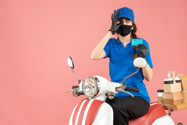 Vooraanzicht van koeriersmeisje met medisch masker en handschoenen zittend op een scooter met een bankkaart die bestellingen aflevert en een brilgebaar maakt op een pastelkleurige perzikachtergrond