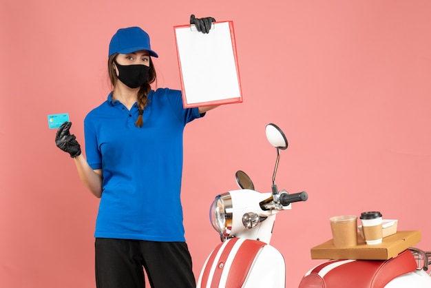 Vooraanzicht van koeriersmeisje met medische maskerhandschoenen die naast motorfiets staan met koffiecake erop met documenten bankkaart op pastel perzikkleurige achtergrond