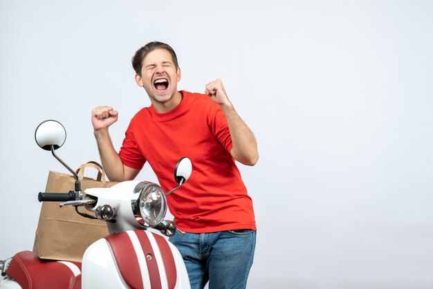 Vooraanzicht van lachende bezorger in rood uniform staande in de buurt van scooter gevoel erg blij op witte achtergrond