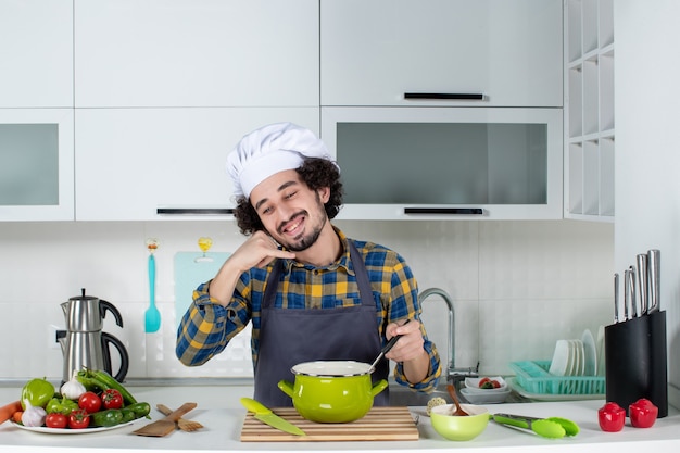 Vooraanzicht van lachende mannelijke chef-kok met verse groenten en het mengen van een maaltijd die me een gebaar maakt in de witte keuken