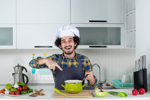 Vooraanzicht van lachende mannelijke chef-kok met verse groenten en wijzende maaltijd in de witte keuken