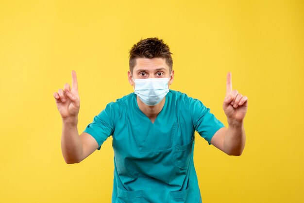 Vooraanzicht van mannelijke arts in medisch kostuum en beschermend masker op gele muur