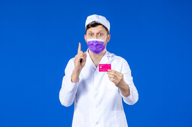 Vooraanzicht van mannelijke arts in medisch kostuum en de creditcard van de maskerholding op blauw