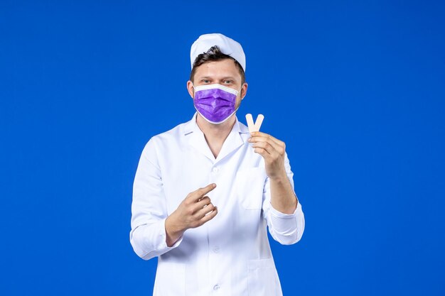 Vooraanzicht van mannelijke arts in medisch kostuum en masker die kleine medische flarden op blauw houden