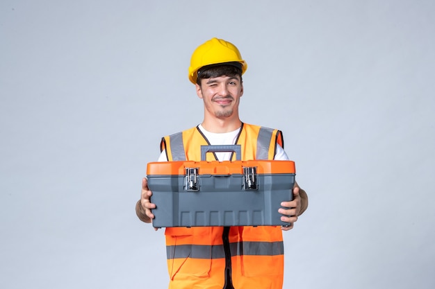 Vooraanzicht van mannelijke bouwer in uniforme en gele helm met gereedschapskoffer op witte muur