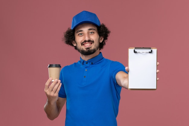 Vooraanzicht van mannelijke koerier in blauw uniform en pet met koffiekopje en blocnote op zijn handen op de roze muur