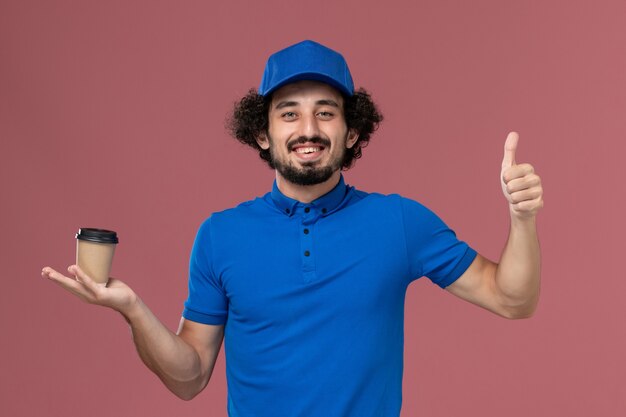 Vooraanzicht van mannelijke koerier in blauw uniform en pet met koffiekopje levering op zijn handen op roze muur