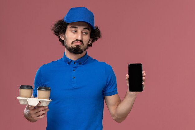 Vooraanzicht van mannelijke koerier in blauw uniform en pet met koffiekopjes voor levering en werktelefoon op zijn handen op de roze muur