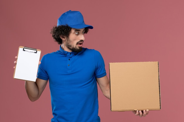 Vooraanzicht van mannelijke koerier in blauwe uniforme pet met blocnote en voedseldoos op zijn handen op roze muur