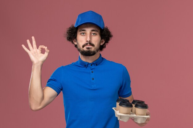 Vooraanzicht van mannelijke koerier in blauwe uniforme pet met koffiekopjes levering op zijn handen op lichtroze muur