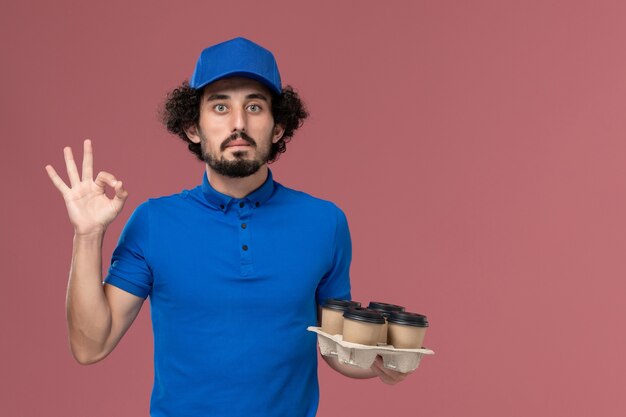 Vooraanzicht van mannelijke koerier in blauwe uniforme pet met koffiekopjes levering op zijn handen op lichtroze muur