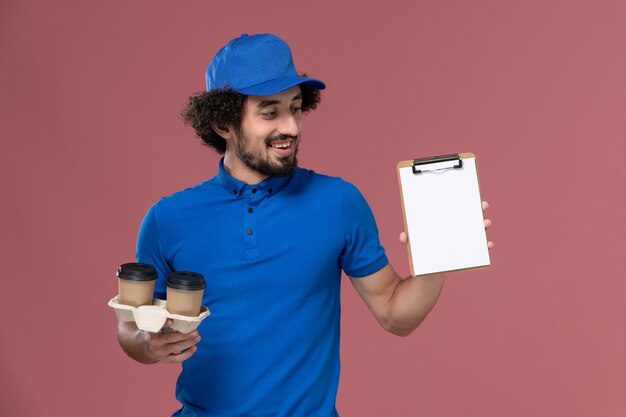 Vooraanzicht van mannelijke koerier in blauwe uniforme pet met koffiekopjes voor levering en blocnote op zijn handen op roze muur