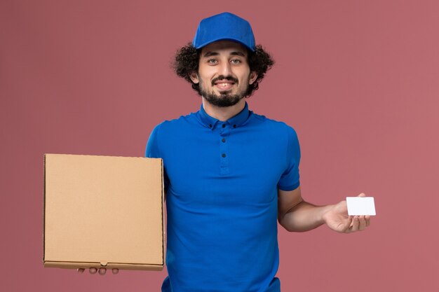 Vooraanzicht van mannelijke koerier in blauwe uniforme pet met voedseldoos en witte plastic kaart op zijn handen op roze muur