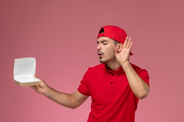 Vooraanzicht van mannelijke koerier in rood uniform en pet die een klein afleverpakket vasthoudt en probeert te horen op de roze muur