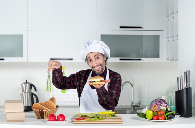 Vooraanzicht van mannelijke kok die hamburger omhoog houdt wijzend op brood op tafel die achter de keukentafel staat