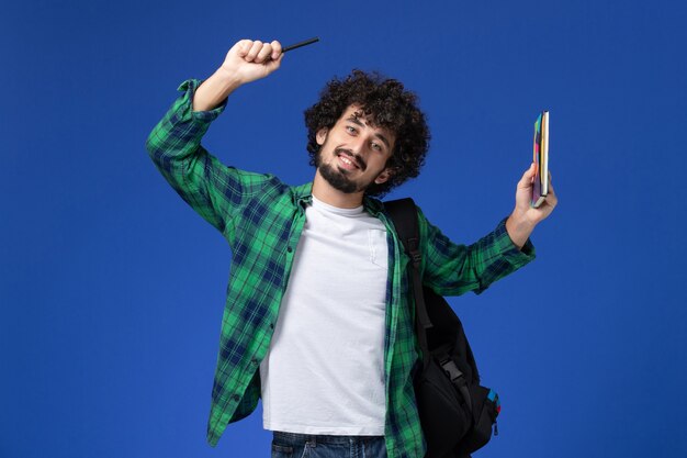 Vooraanzicht van mannelijke student die het zwarte voorbeeldenboek van de rugzakholding en pen met glimlach op blauwe muur dragen