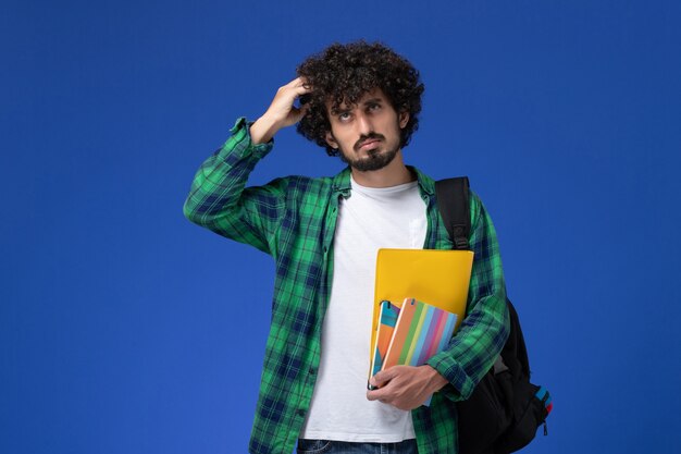 Vooraanzicht van mannelijke student die zwarte rugzak draagt die voorbeeldenboeken en dossiers houdt die aan de blauwe muur denken