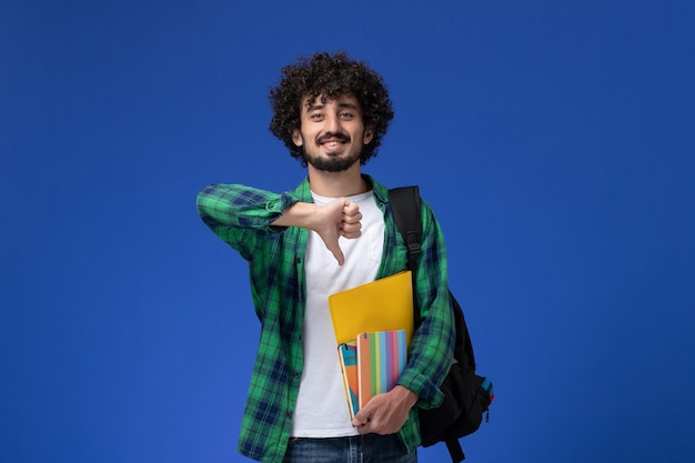 Vooraanzicht van mannelijke student die zwarte rugzak draagt die voorbeeldenboeken en dossiers op de blauwe muur houdt