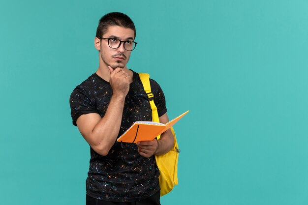 Vooraanzicht van mannelijke student in donkere t-shirt gele rugzak houden en voorbeeldenboek lezen denken op blauwe muur