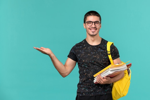 Vooraanzicht van mannelijke student in donkere t-shirt gele rugzak met bestanden en boeken glimlachen op lichtblauwe muur