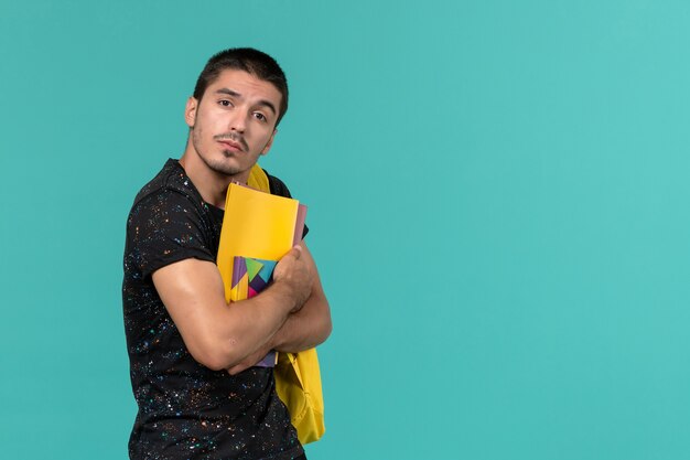 Vooraanzicht van mannelijke student in donkere t-shirt gele rugzak met bestanden en voorbeeldenboek op de lichtblauwe muur