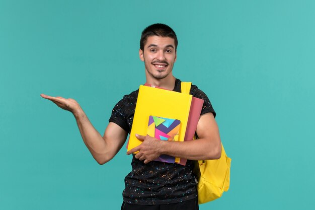 Vooraanzicht van mannelijke student in donkere t-shirt gele rugzak met bestanden en voorbeeldenboek op de lichtblauwe muur