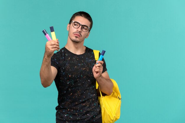 Vooraanzicht van mannelijke student in donkere t-shirt gele rugzak met gekleurde viltstiften op blauwe muur