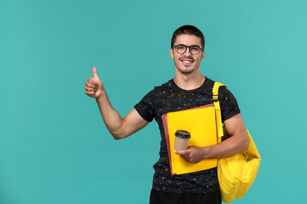 Vooraanzicht van mannelijke student in donkere t-shirt gele rugzak met verschillende bestanden en koffie op de blauwe muur