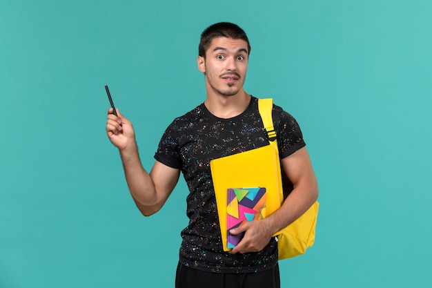 Vooraanzicht van mannelijke student in donkere t-shirt gele rugzak met voorbeeldenboek pen en bestanden op de lichtblauwe muur