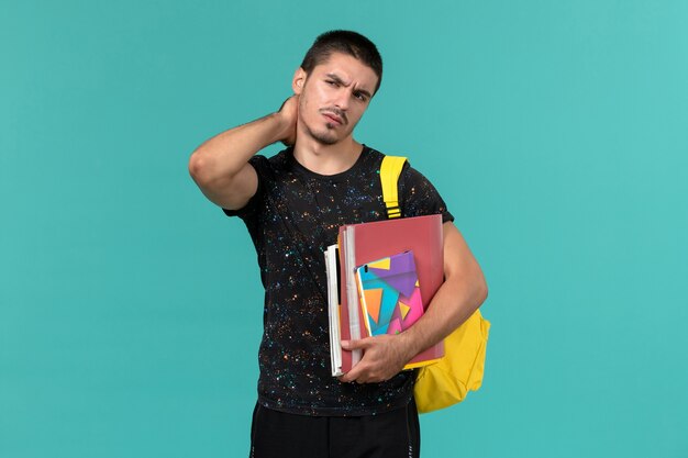 Vooraanzicht van mannelijke student in donkere t-shirt met gele rugzak met voorbeeldenboek en bestanden met nekpijn op blauwe muur