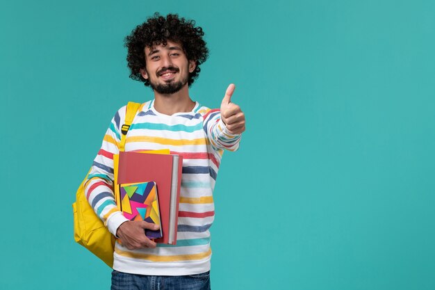 Vooraanzicht van mannelijke student in gekleurd gestreept overhemd die gele rugzak dragen die dossiers en voorbeeldenboeken houden die op blauwe muur glimlachen