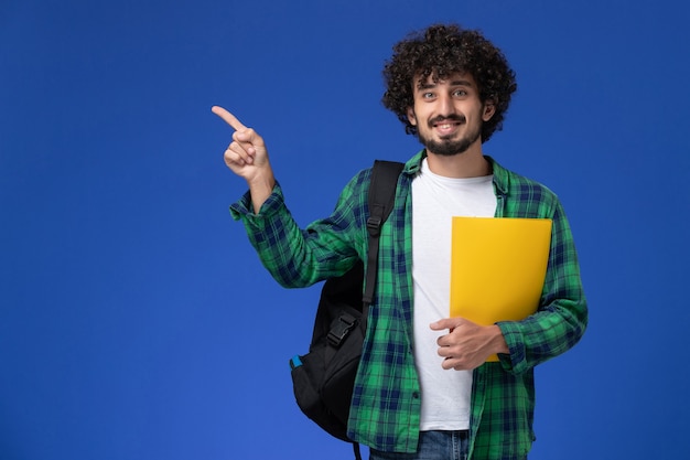 Vooraanzicht van mannelijke student in groen geruit overhemd die zwarte rugzak draagt en dossiers houdt die op de blauwe muur glimlachen