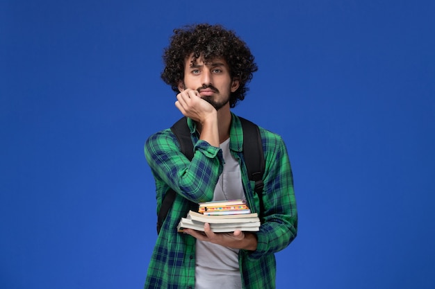Vooraanzicht van mannelijke student in groen geruit overhemd met zwarte rugzak die voorbeeldenboeken en dossiers houden die op blauwe muur denken