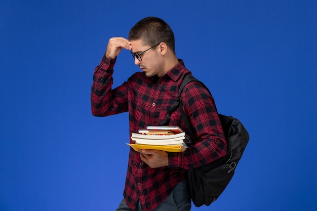 Vooraanzicht van mannelijke student in rood geruit hemd met rugzak met voorbeeldenboeken en voorbeeldenboeken op lichtblauwe muur