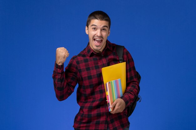 Vooraanzicht van mannelijke student in rood geruit overhemd met de dossiers van de rugzakholding en voorbeeldenboeken die zich op blauwe muur verheugen