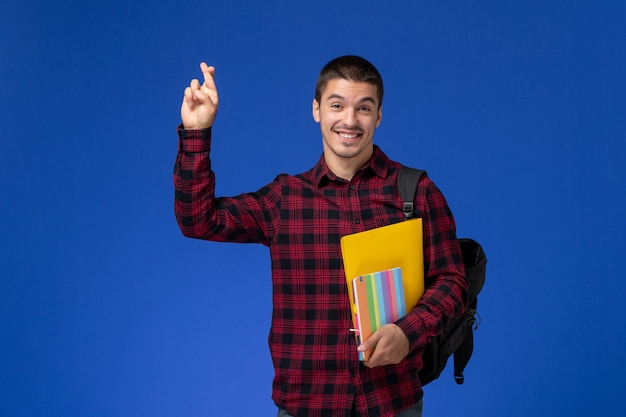 Vooraanzicht van mannelijke student in rood geruit overhemd met rugzak met bestanden en voorbeeldenboeken op de blauwe muur