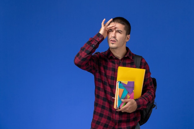 Vooraanzicht van mannelijke student in rood geruit overhemd met rugzak met bestanden en voorbeeldenboeken op lichtblauwe muur